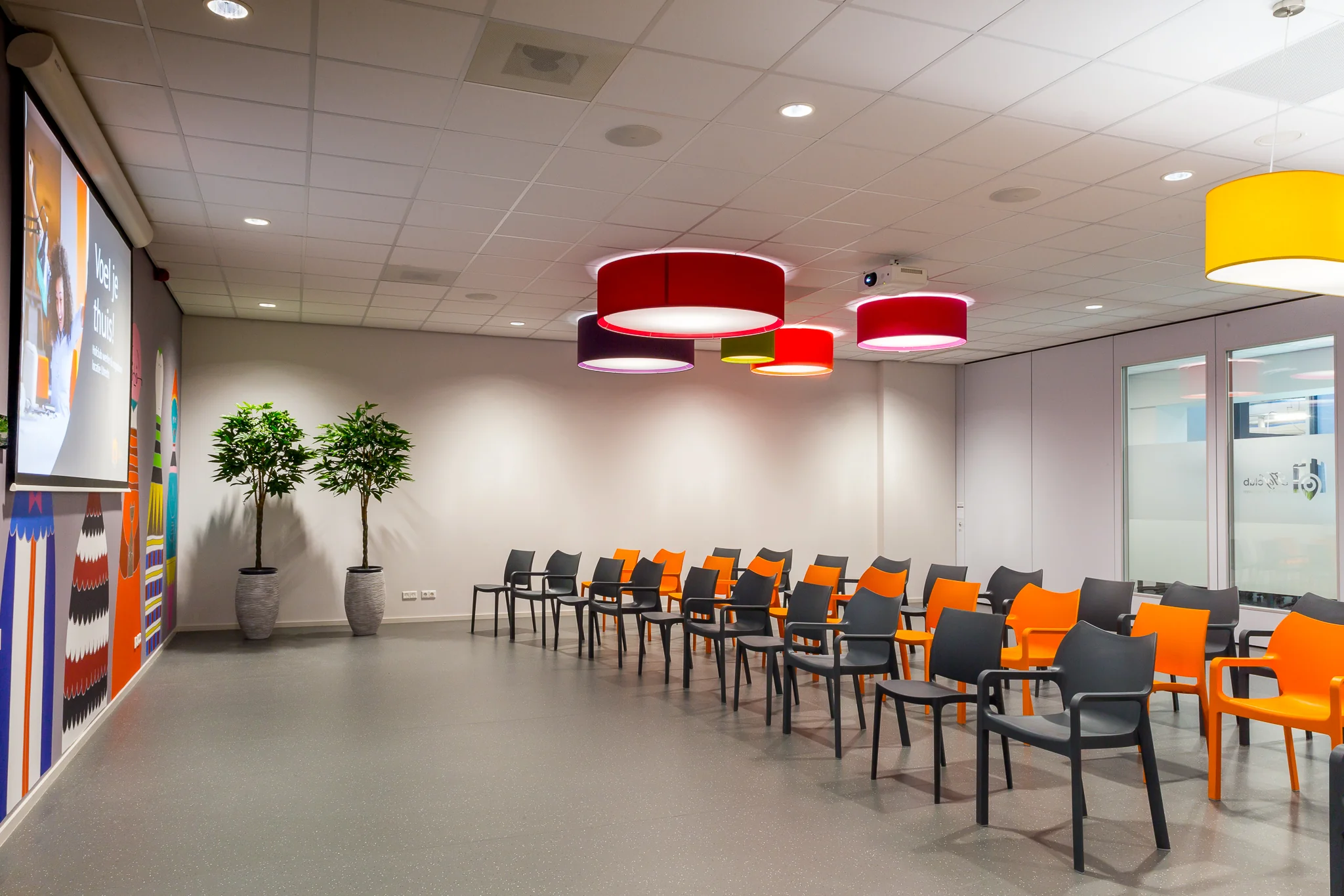 Kleurrijke wachtruimte met zwarte en oranje stoelen, grote ronde plafondlampen en een projectiescherm aan de Niels Bohrweg.